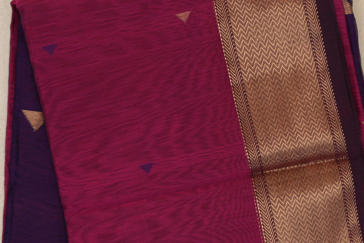 Close-up of a folded deep rani pink Maheshwari silk cotton saree with small triangle motifs and a wide gold zigzag border beside deep purple fabric.