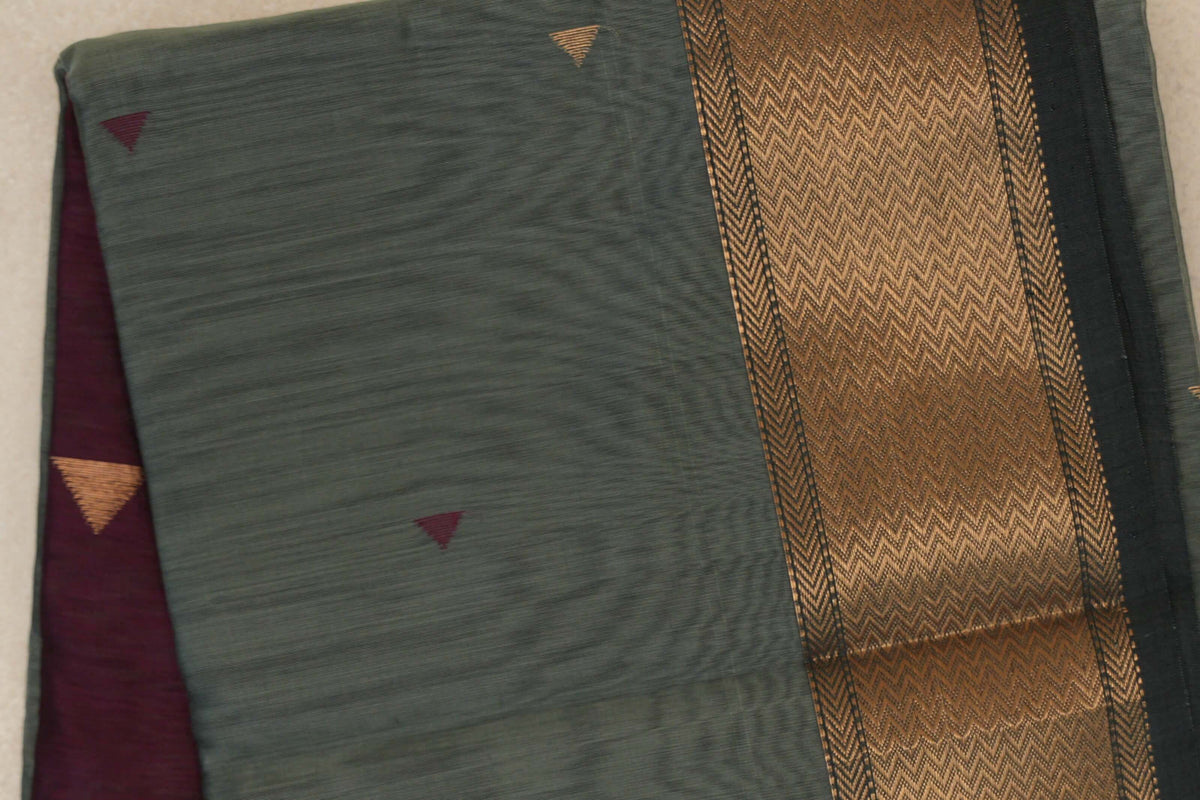 Folded Maheshwari silk cotton saree showing a sage green body with small triangular motifs, a gold zigzag border and a glimpse of deep wine maroon fabric.