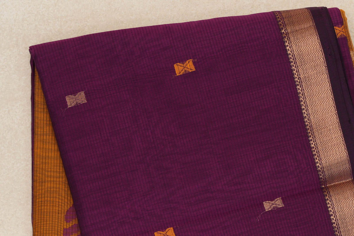 Folded deep magenta Maheshwari silk cotton saree with small gold and mustard accents, a patterned gold border and mustard orange fabric peeking beneath.