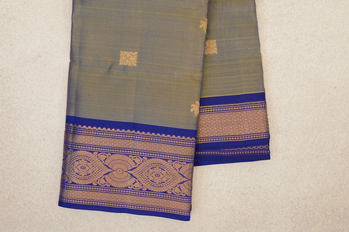 Folded light weight Kanchipuram silk saree with a muted yellow-violet body, small gold squares and a deep royal blue border with ornate gold patterns.