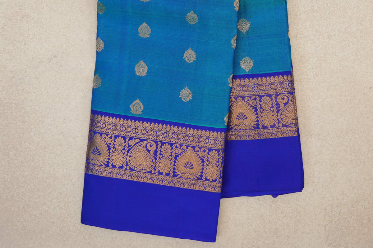 Folded peacock blue Kanchipuram silk saree with scattered gold-toned motifs and royal blue borders featuring paisley and floral patterns.