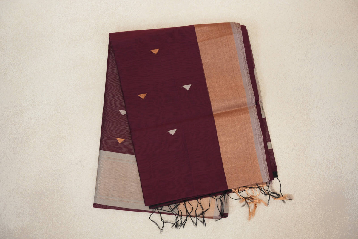 Folded deep wine maroon Maheshwari silk cotton saree with small triangular motifs, a broad warm-toned border, silver grey selvedge and tassels.