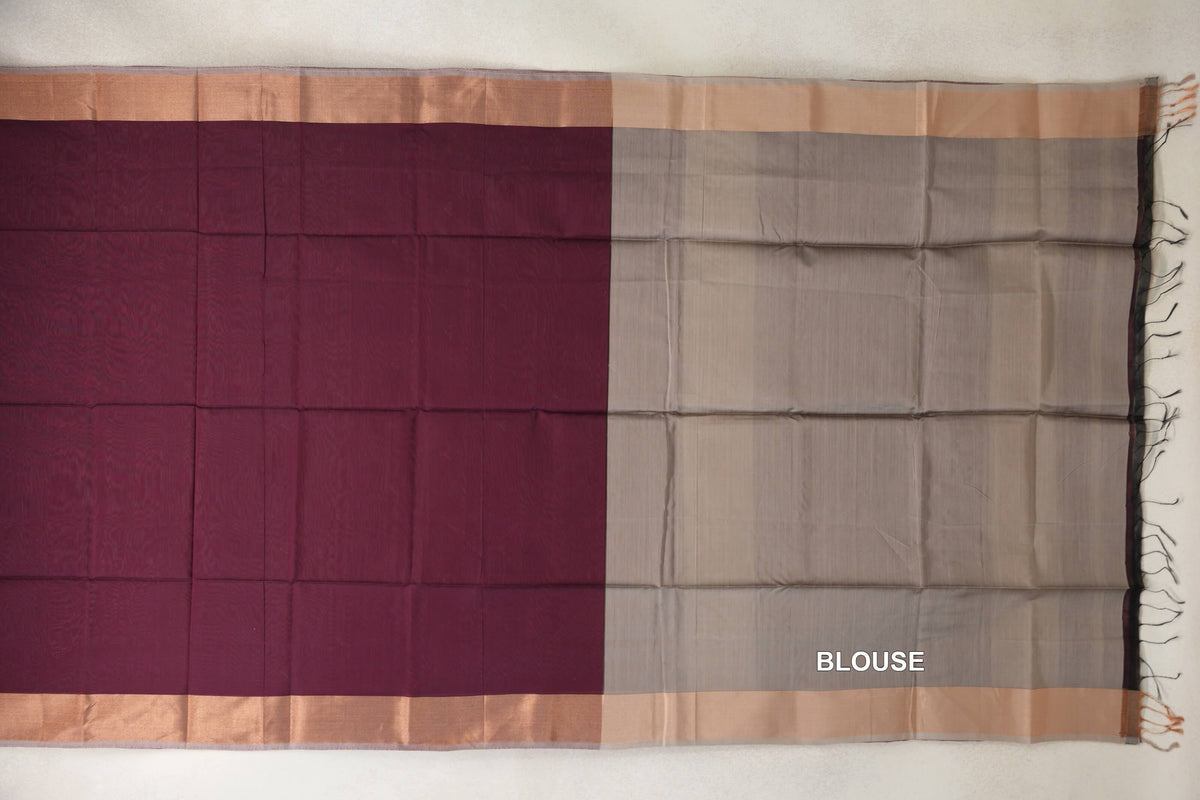 Maheshwari silk cotton saree laid flat, showing a deep wine maroon body beside a dark beige blouse piece, with broad borders, silver grey edging and tassels.
