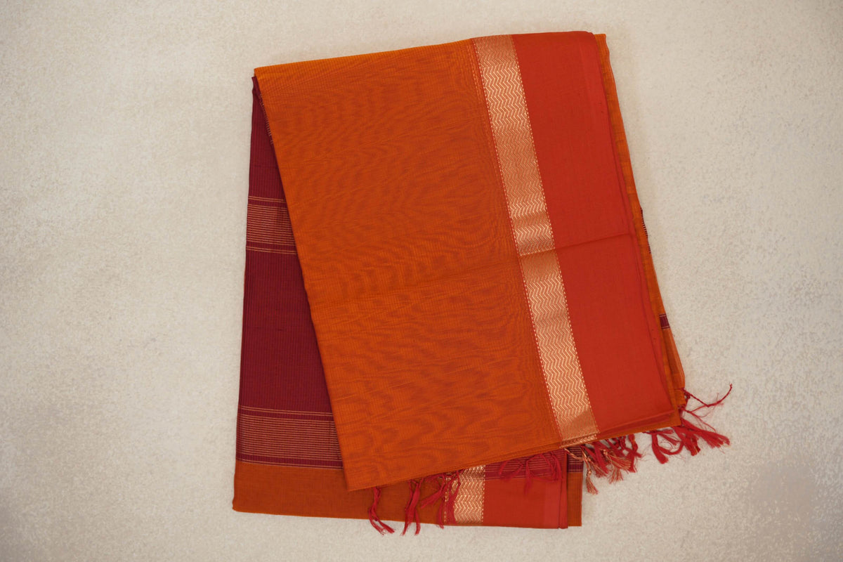 Folded Maheshwari silk cotton saree with a reddish orange body, orangish red border, gold zigzag band and a striped reddish maroon section with tassels.
