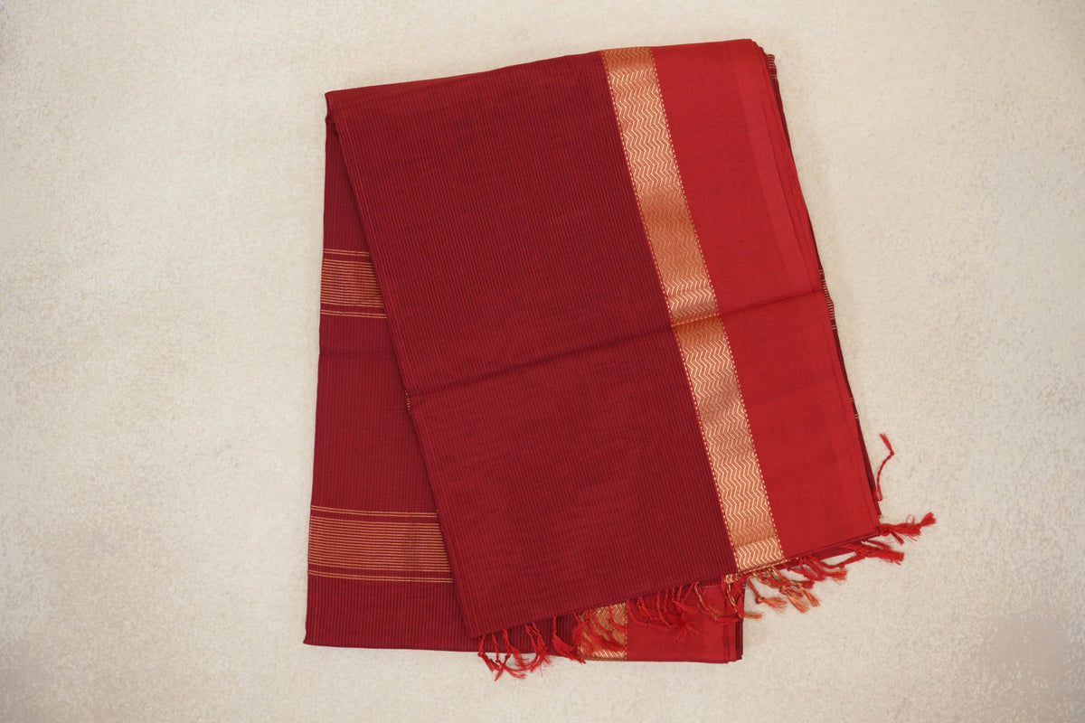 Folded deep red Maheshwari silk cotton saree with fine stripes, a gold-toned zigzag border and tassels along the edge.