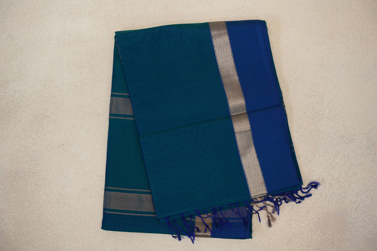 Folded peacock blue Maheshwari silk cotton saree with a deep blue panel, silver zigzag border, fine stripes and tassels.
