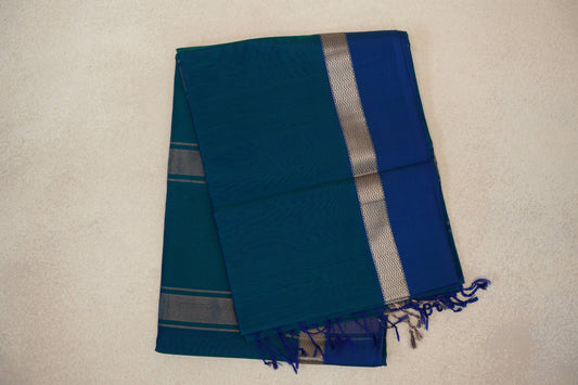 Folded peacock blue Maheshwari silk cotton saree with a deep blue panel, silver zigzag border, fine stripes and tassels.