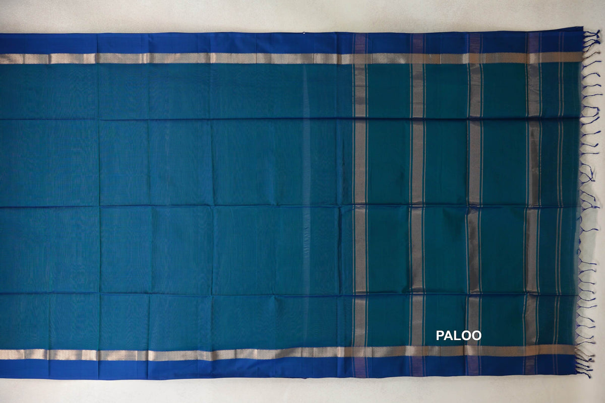 Peacock blue Maheshwari silk cotton saree laid flat, showing a plain body, silver-toned border bands and a striped pallu with blue tassels.