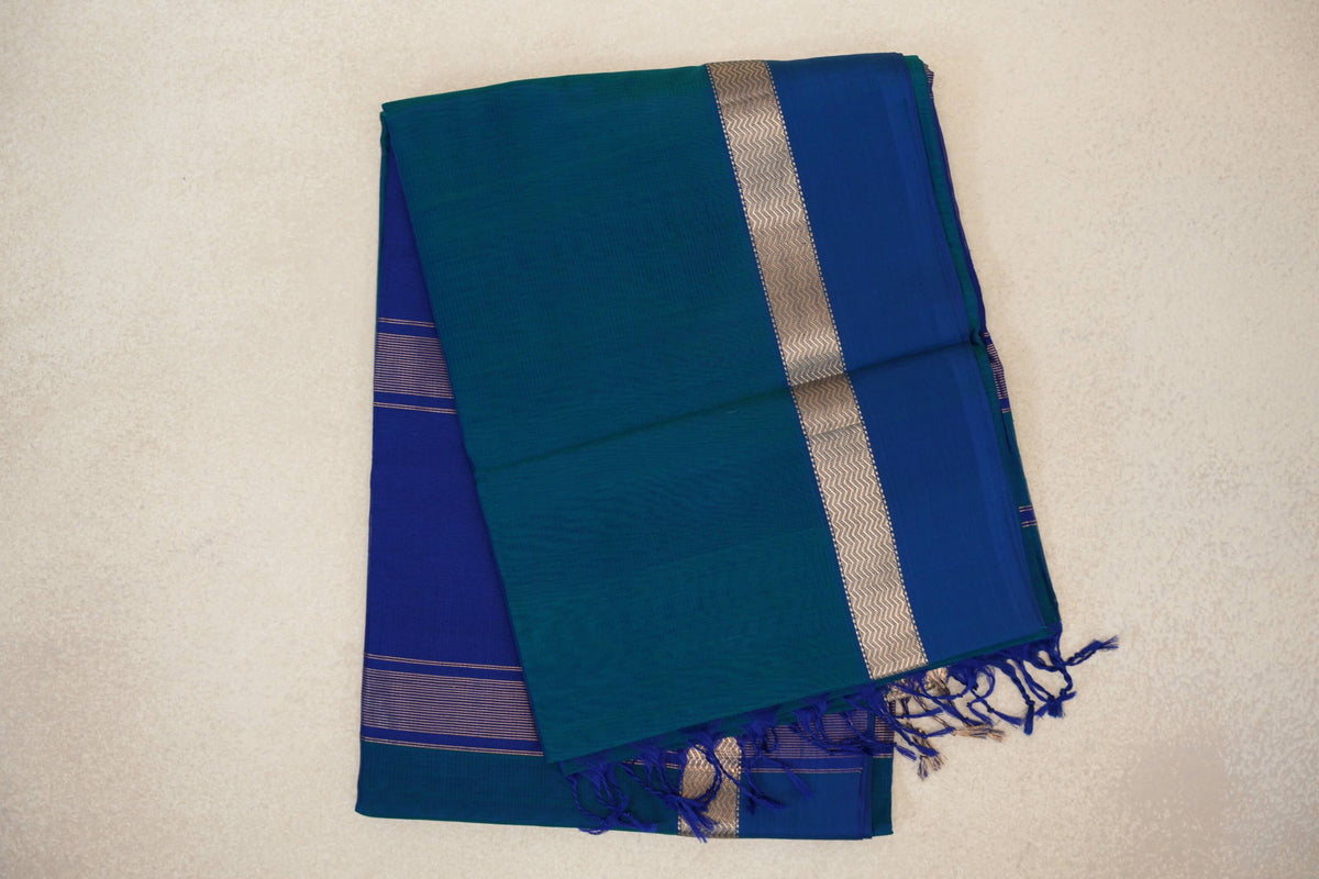 Folded peacock blue Maheshwari silk cotton saree with a deep blue border, metallic zigzag trim, striped pallu and blue tassels.