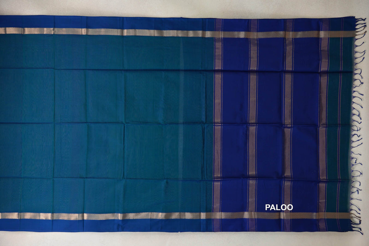 Maheshwari silk cotton saree laid flat, showing a plain peacock blue body, deep blue striped pallu, silver-toned borders and tassels.