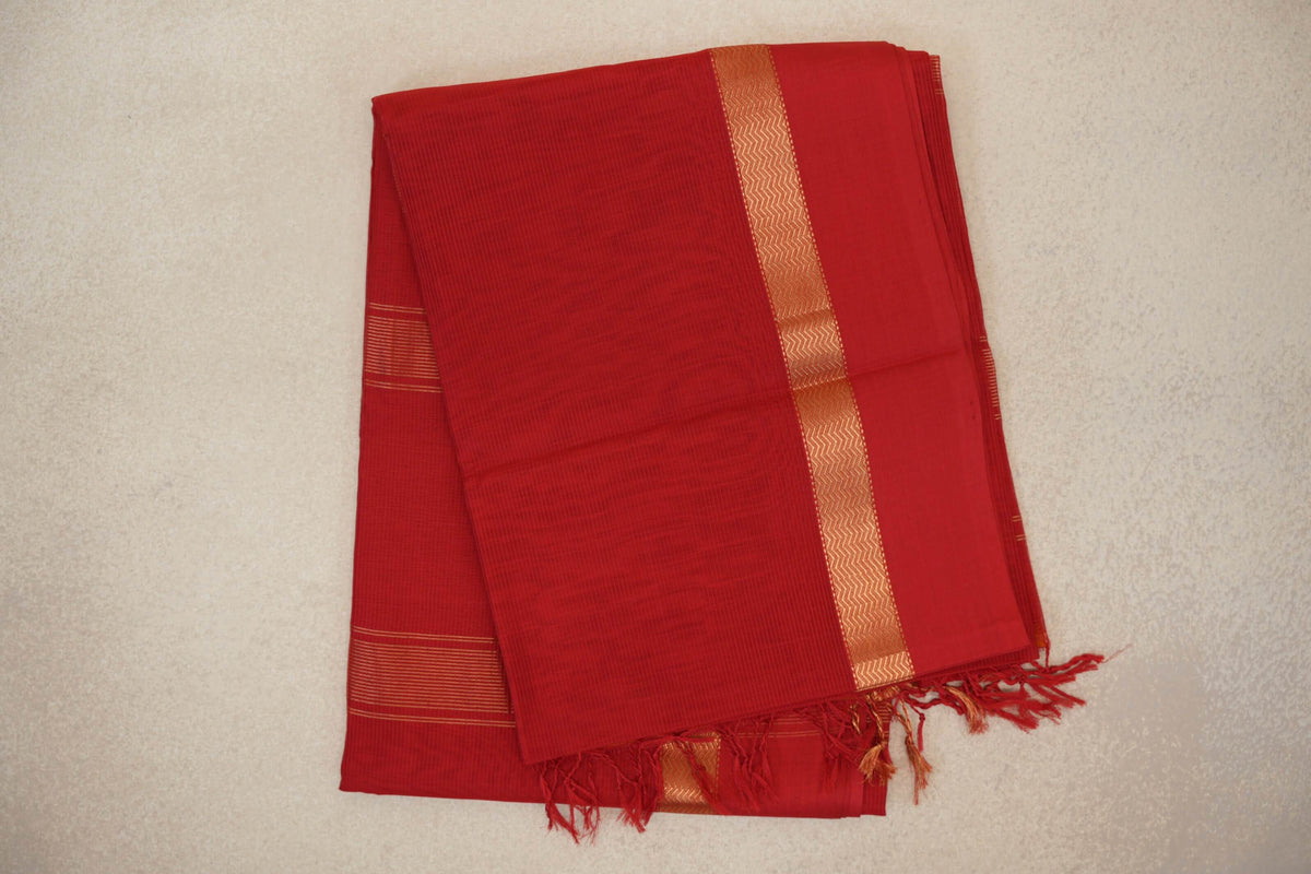 Folded vermilion red Maheshwari silk cotton saree with a fine checked weave, gold zigzag border, narrow stripes and red tassels.
