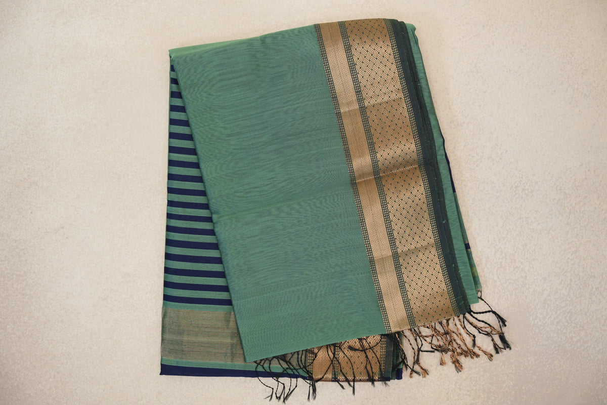 Folded sea green Maheshwari silk cotton saree with a dark sea green and gold-toned patterned border; navy-striped pallu and tassels peek out beneath.