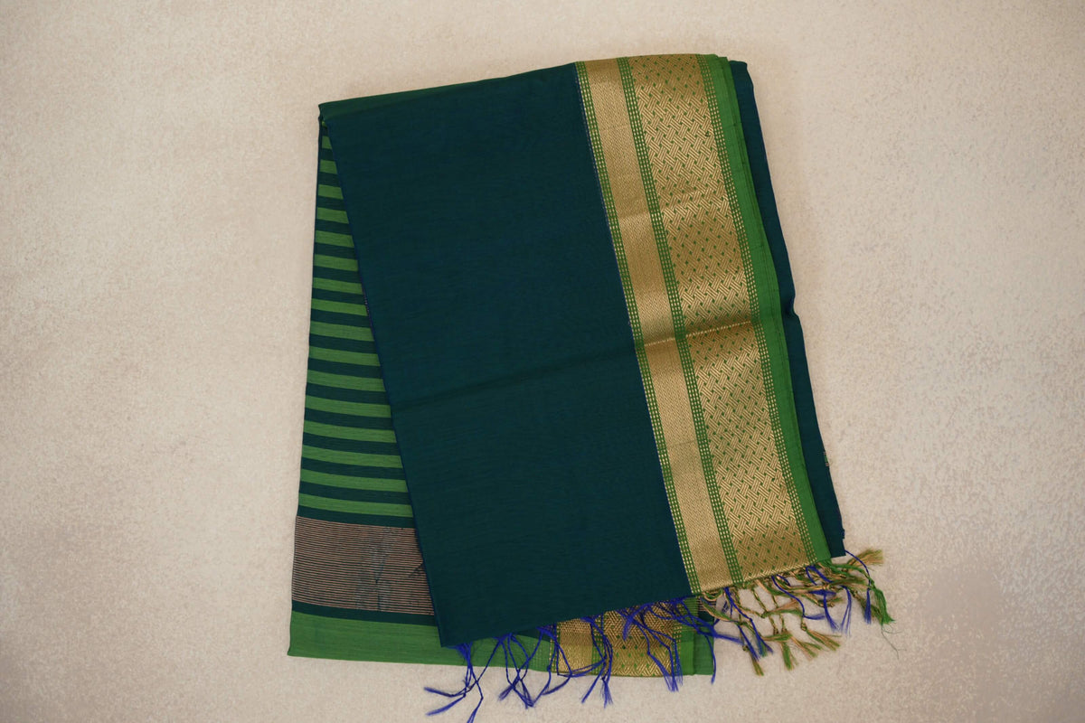 Folded Maheshwari silk cotton saree with a peacock blue body, leaf green and gold-toned patterned border, striped pallu and tassels.