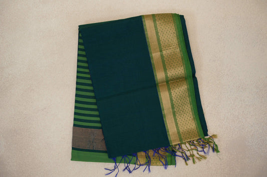 Folded Maheshwari silk cotton saree with a peacock blue body, leaf green and gold-toned patterned border, striped pallu and tassels.