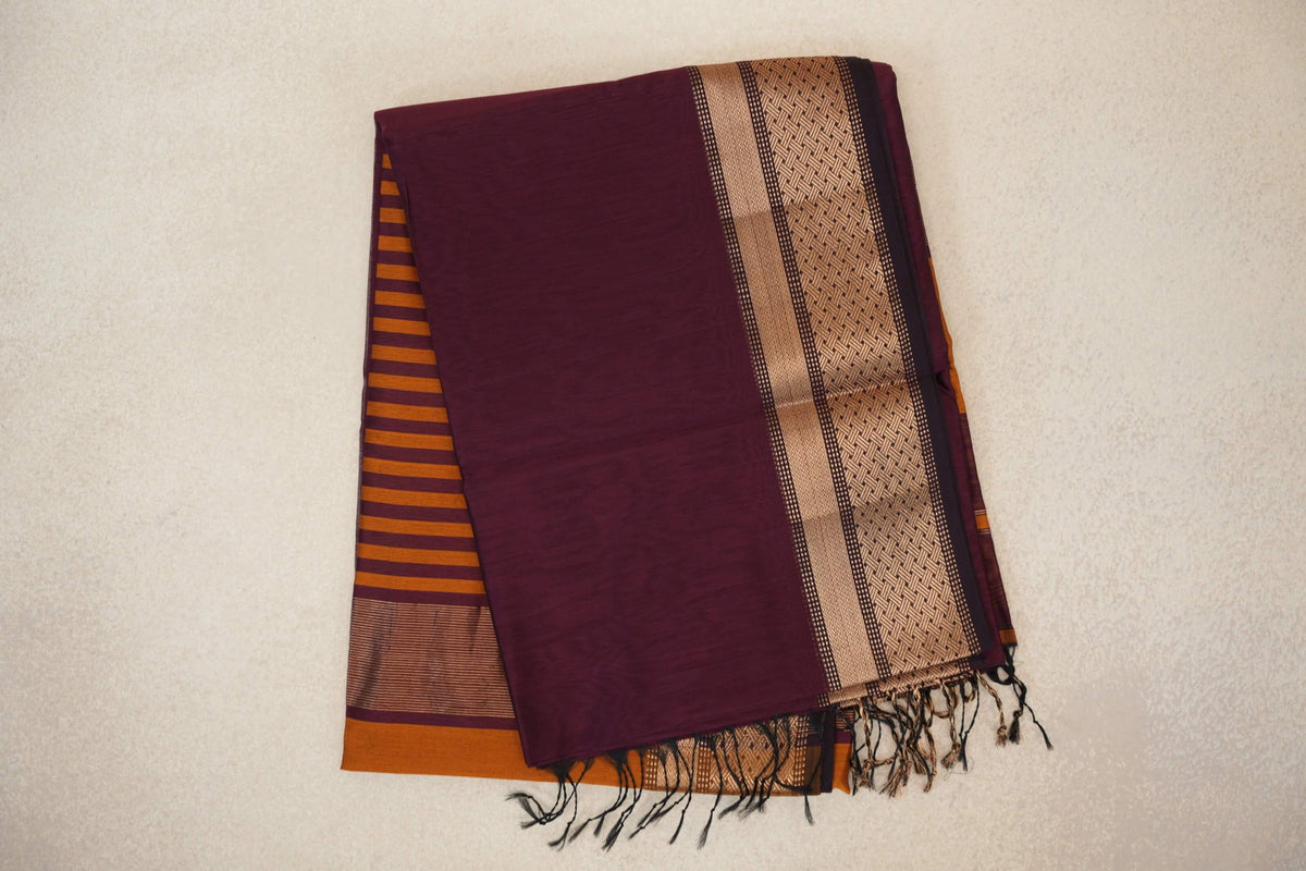 Folded deep plum Maheshwari silk cotton saree with a gold-toned geometric border, deep orange striped pallu peeking out and tassels.