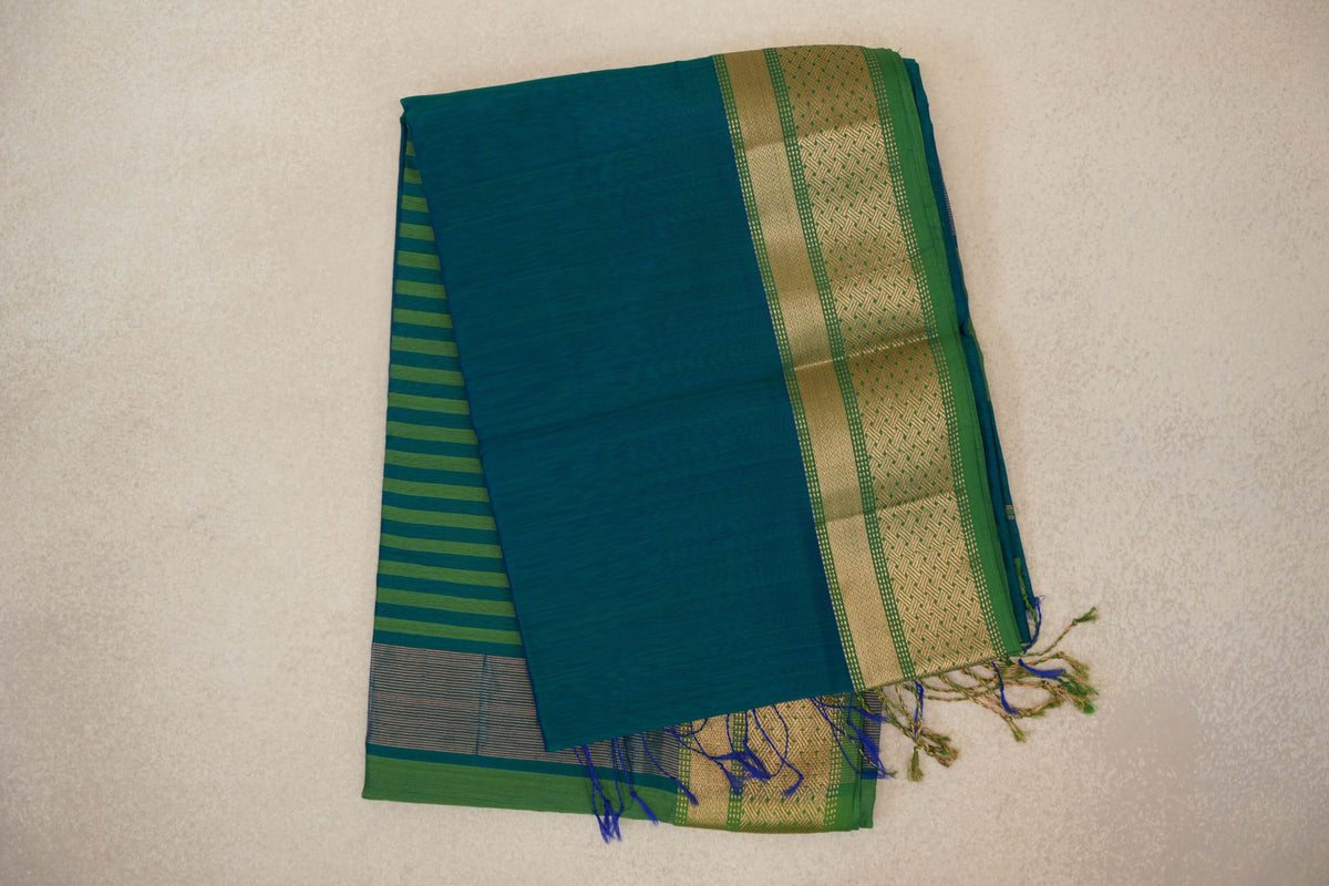 Folded Maheshwari silk cotton saree with a peacock green body, leaf green border with gold geometric detailing, striped fabric beneath and tassels.