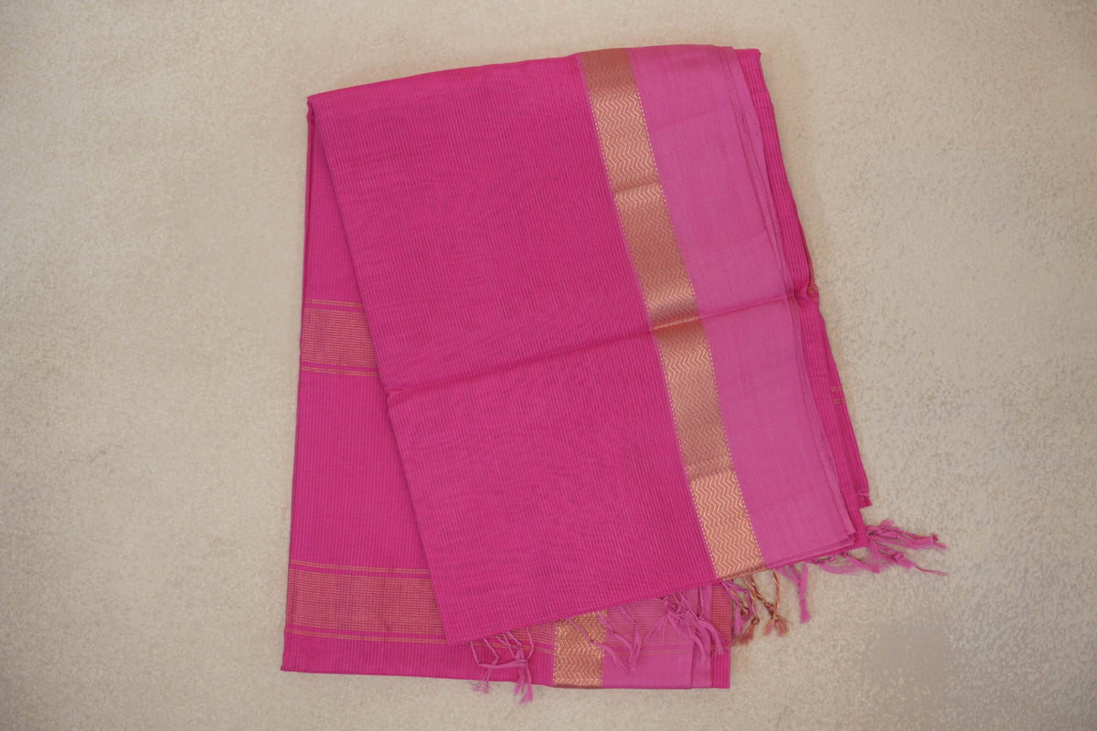 Folded bright rani pink Maheshwari silk cotton saree with fine stripes, a pink border edged in gold zigzags, and tassels.