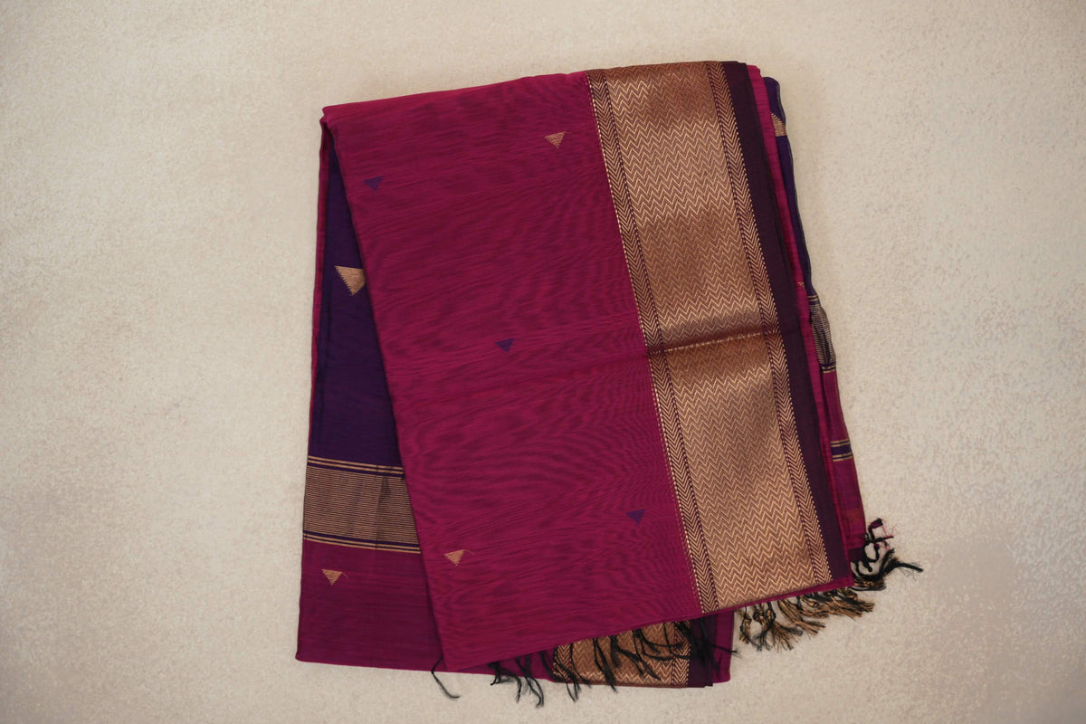 Folded deep rani pink Maheshwari silk cotton saree with small triangular accents, a wide gold chevron-patterned border, deep purple folds and tassels.
