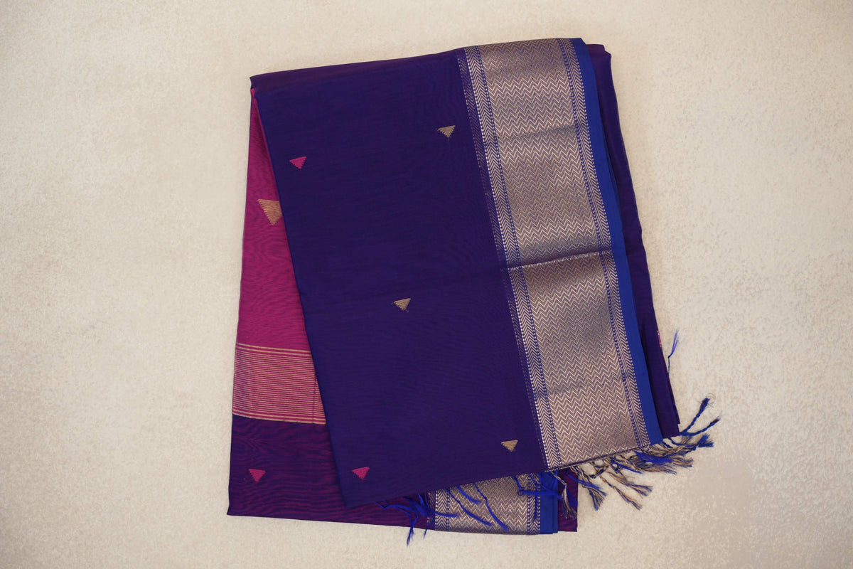 Folded deep violet Maheshwari silk cotton saree with small triangle motifs, a gold-toned chevron border, blue tassels and deep rani pink fabric beneath.
