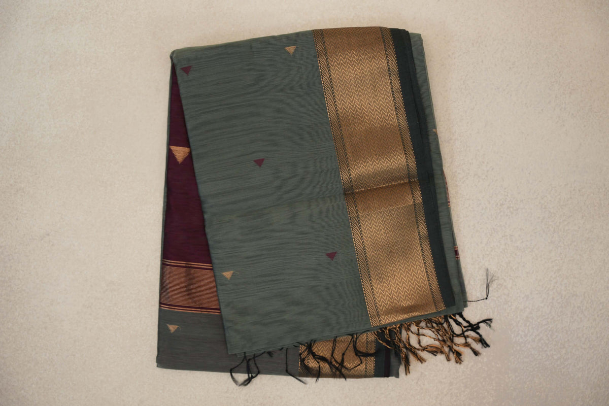 Folded Maheshwari silk cotton saree in sage green with small triangle motifs, a gold chevron-patterned border, maroon fabric peeking out and tassels.