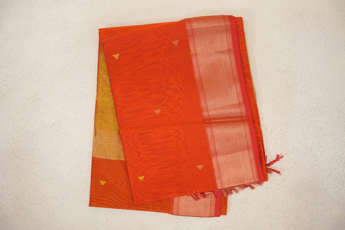 Folded Maheshwari silk cotton saree with a reddish orange body, small triangle motifs, an orangish red chevron border and lime yellow fabric peeking out.