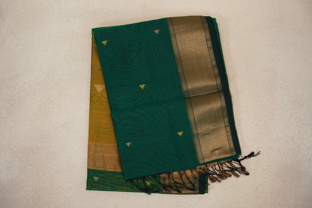Folded bottle green Maheshwari silk cotton saree with small gold accents, a wide chevron-patterned border, mustard yellow fabric beneath and tassels.