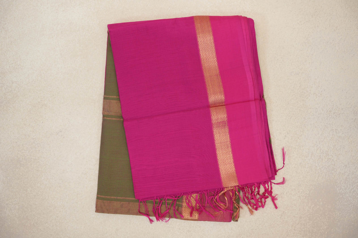 Folded rani pink Maheshwari silk cotton saree with a gold-toned zigzag border, pink tassels and an olive green section visible beneath.