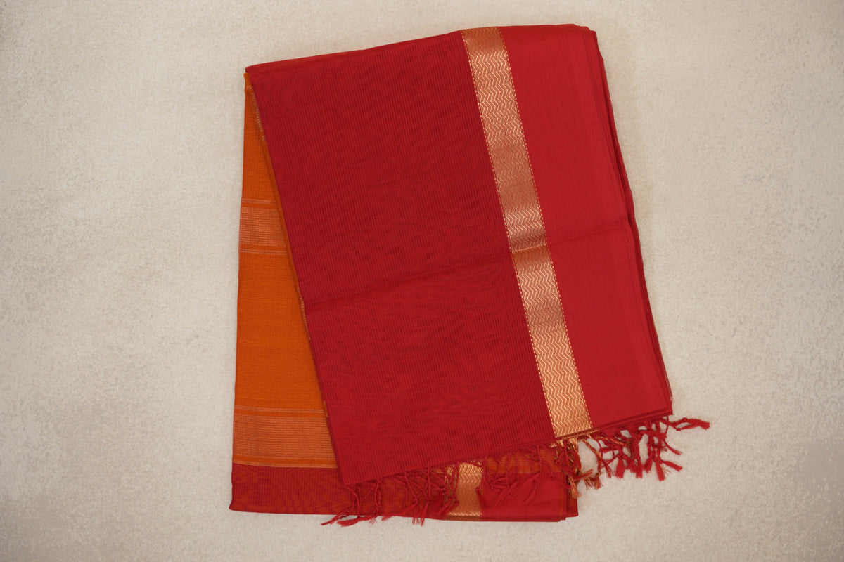 Folded vermilion red Maheshwari silk cotton saree with a gold-toned zigzag border and tassels; deep orange fabric shows beneath.