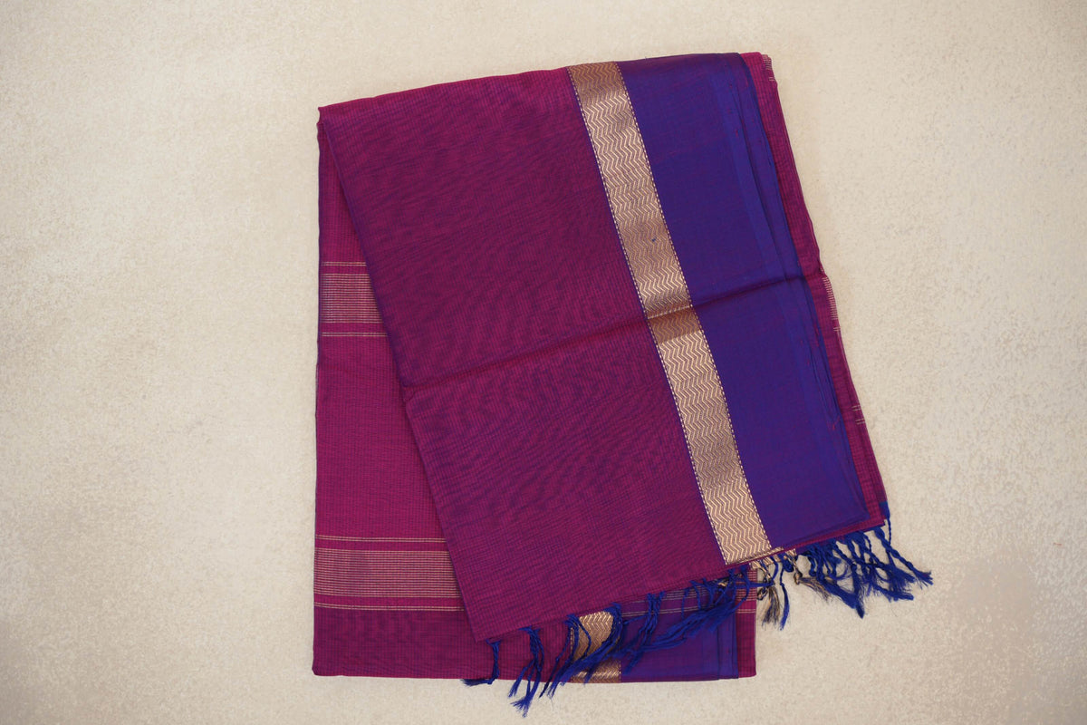 Folded deep magenta Maheshwari silk cotton saree with a violet purple border, gold-toned zigzag band and blue tassels.