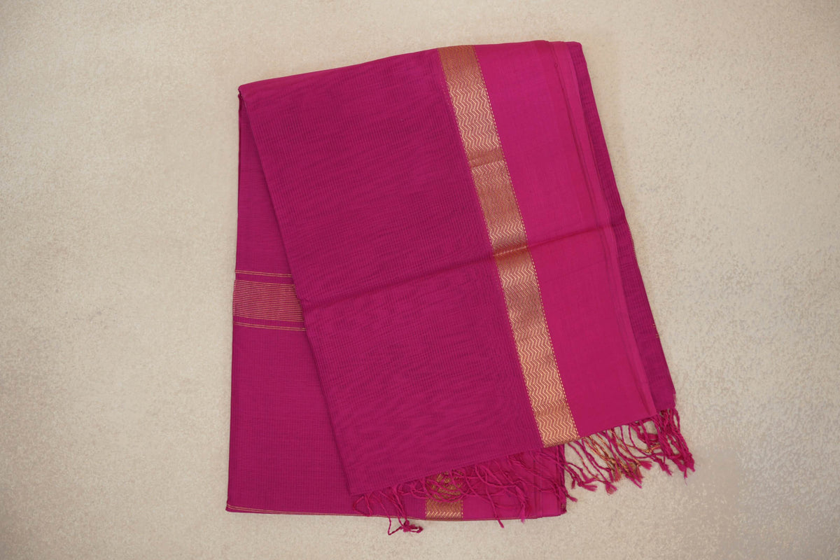 Folded bright rani pink Maheshwari silk cotton saree with a subtle checked weave, gold-toned zigzag border and fringed end.