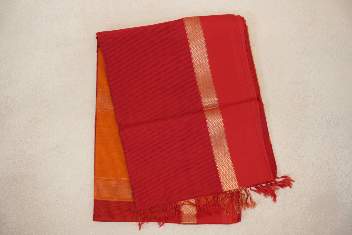 Folded vermilion red Maheshwari silk cotton saree with fine stripes, a gold chevron band and tassels, layered over deep orange fabric.