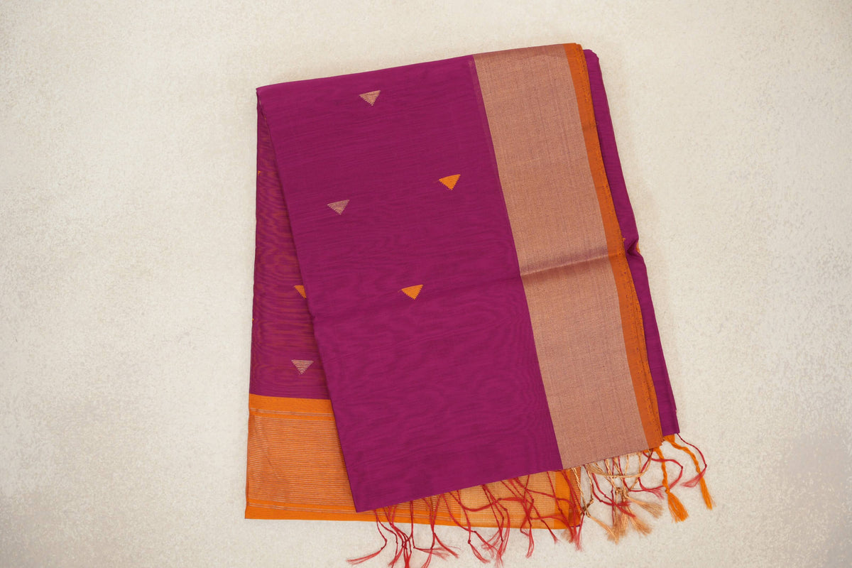 Folded deep magenta Maheshwari silk cotton saree with small triangular motifs, a broad shimmering border, orange selvedge and tassels.