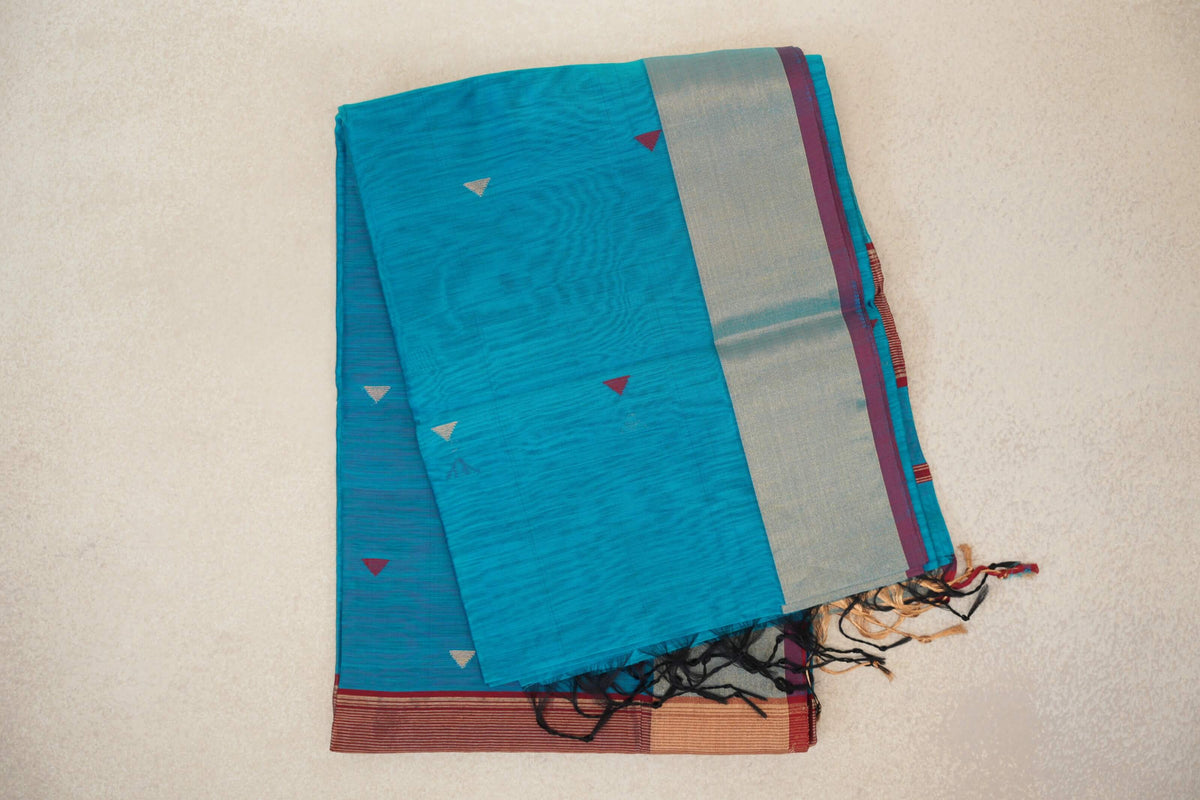 Folded bright turquoise blue Maheshwari silk cotton saree with small triangular motifs, a broad silver-toned border edged in deep red, and tassels.