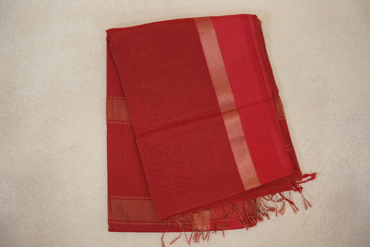 Folded tomato red Maheshwari silk cotton saree with fine gold stripes, a narrow patterned border, a plain red panel and tassels.