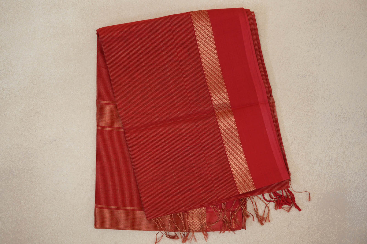 Folded red Maheshwari silk cotton saree with fine light stripes, a broad red border, a narrow geometric band and tassels.
