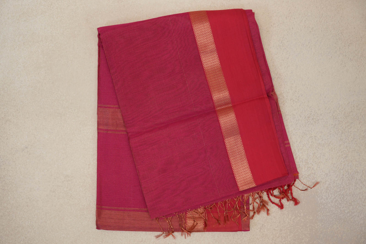 Folded rani pink Maheshwari silk cotton saree with fine stripes, a patterned border, a plain pink panel and tasselled ends.