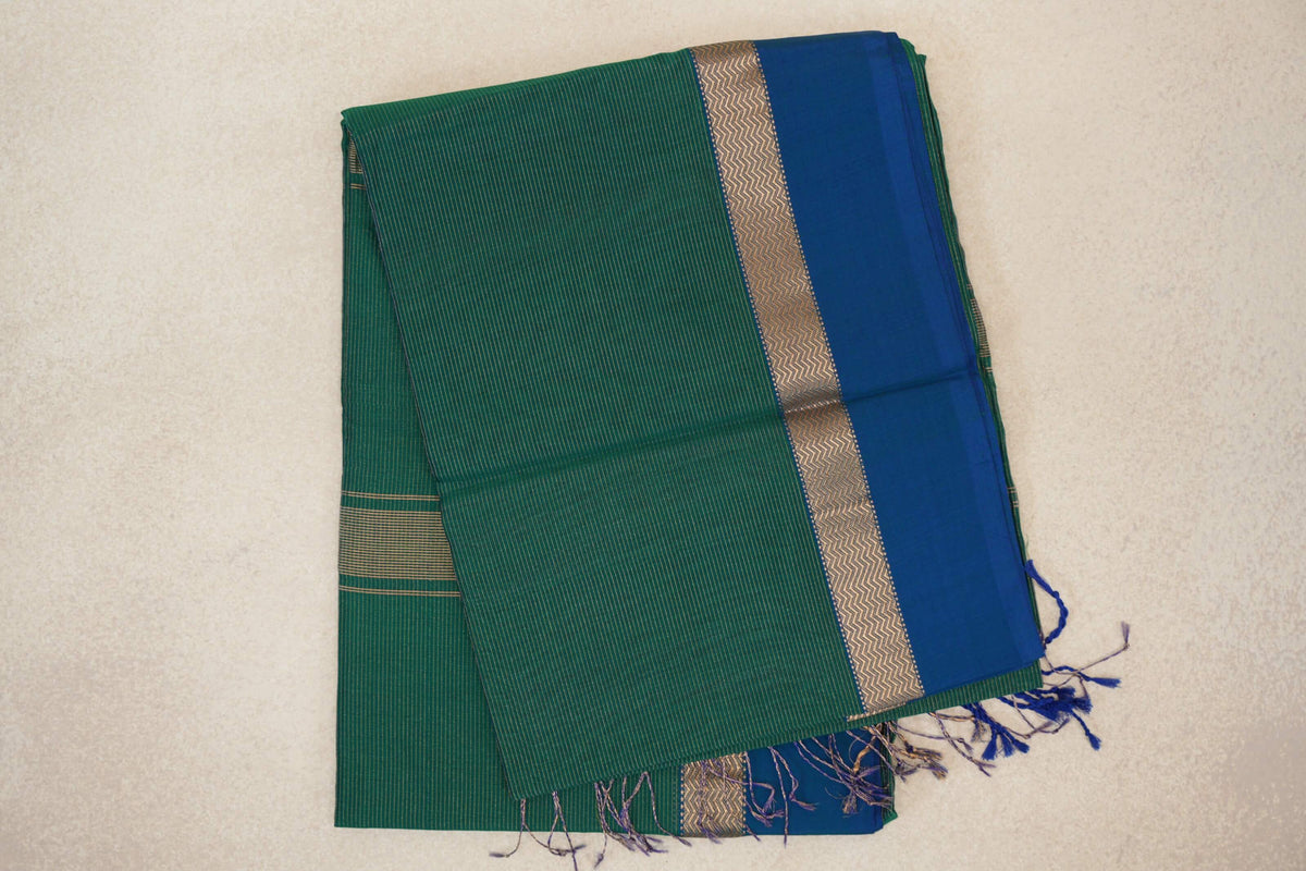 Folded bottle green Maheshwari silk cotton saree with fine stripes, a peacock blue border, chevron-patterned trim and loose tassels.