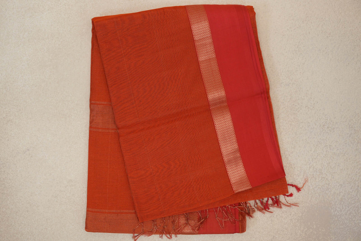 Folded rust orange Maheshwari silk cotton saree with fine stripes, a wide red border edged in zigzags, and tassels at one end.