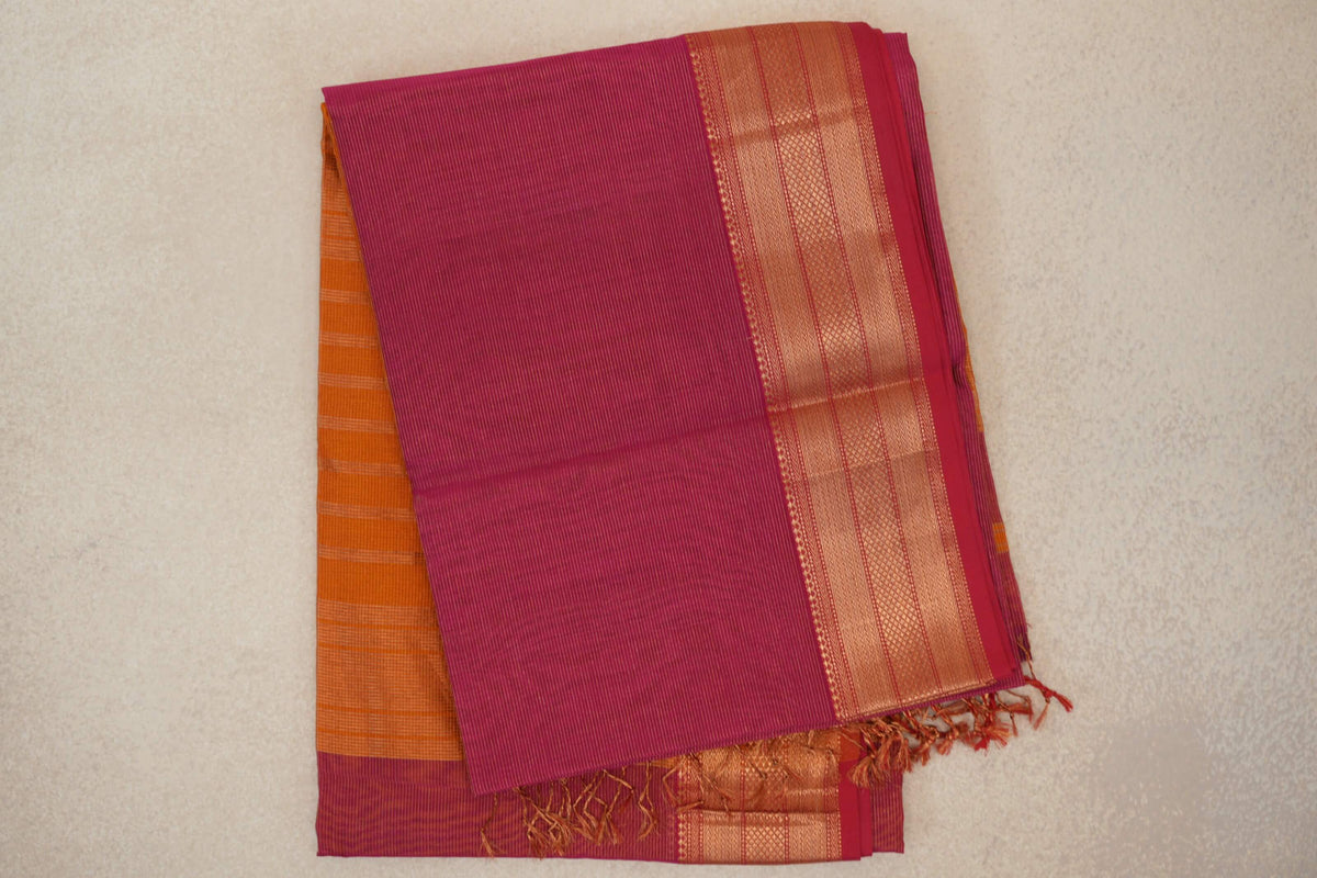 Folded rani pink Maheshwari silk cotton saree with fine stripes, a broad gold-patterned border, orange fabric peeking beneath and tassels.