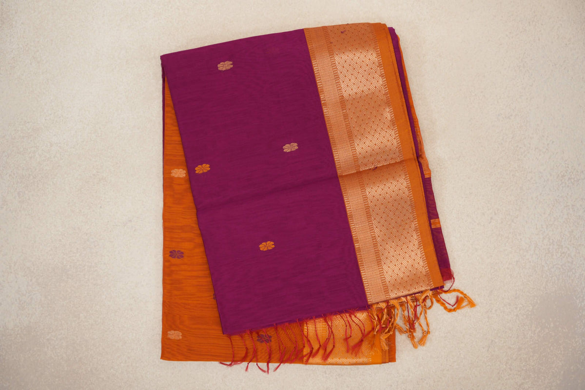 Folded dark magenta Maheshwari silk cotton saree with small floral motifs, a patterned deep orange border and tassels.