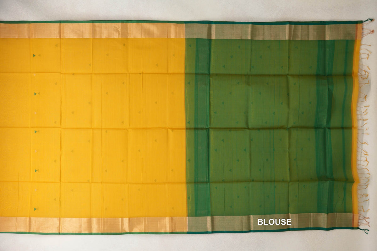 Maheshwari silk cotton saree laid flat, with a turmeric yellow body, small triangular accents, green-edged borders and a bottle green blouse piece at right.