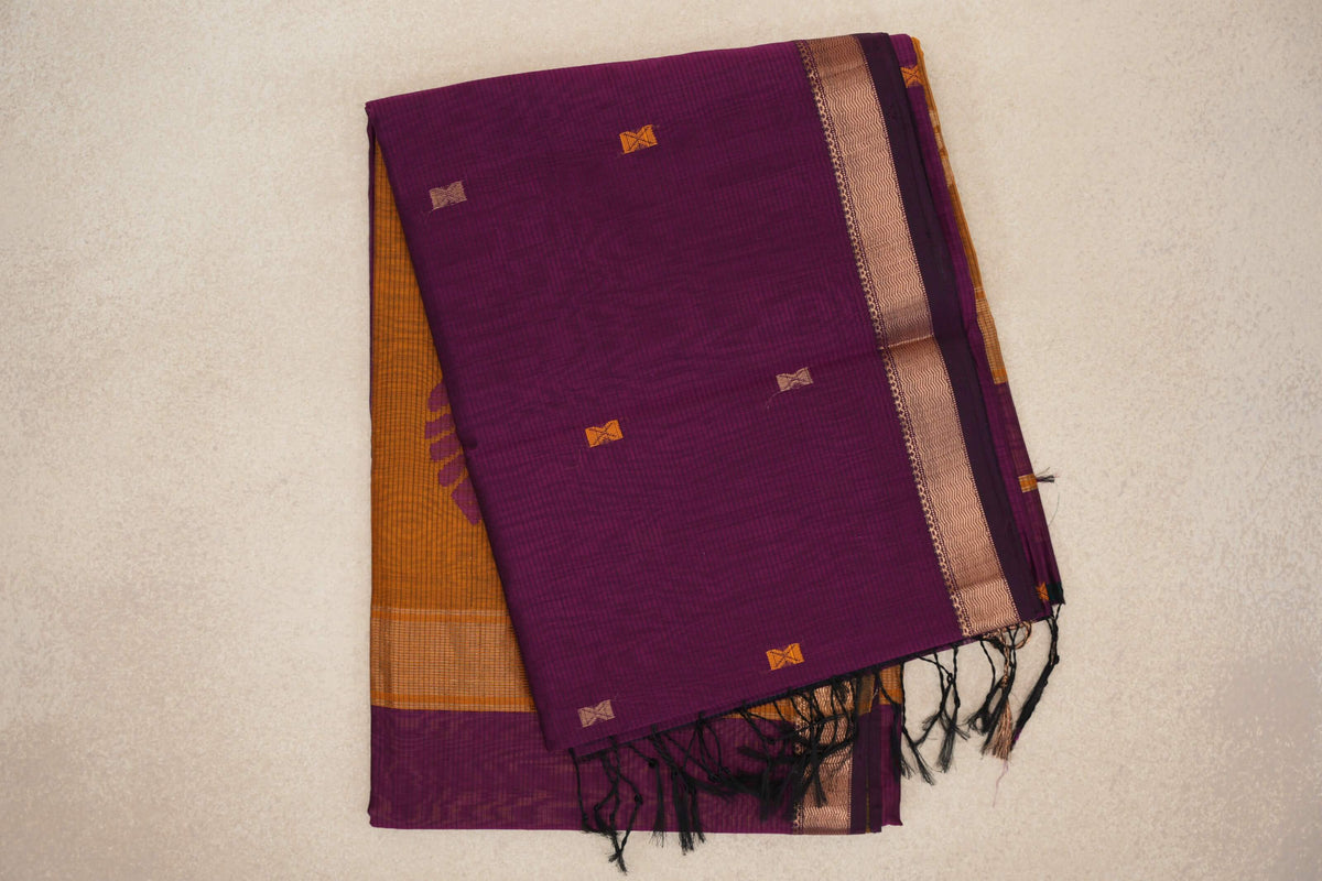 Folded deep magenta Maheshwari silk cotton saree with small woven accents, a patterned gold-toned border, black tassels and mustard orange fabric beneath.
