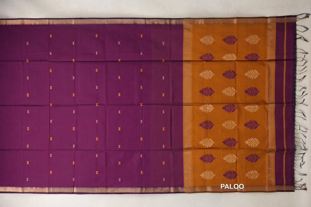 Maheshwari silk cotton saree laid flat, with a deep magenta body, small scattered motifs, and a deep mustard orange pallu with leaf designs and tassels.