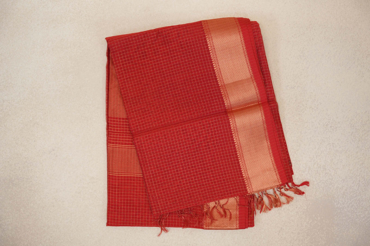 Folded tomato red Maheshwari silk cotton saree with fine gold checks, a broad gold zari border and red tassels.