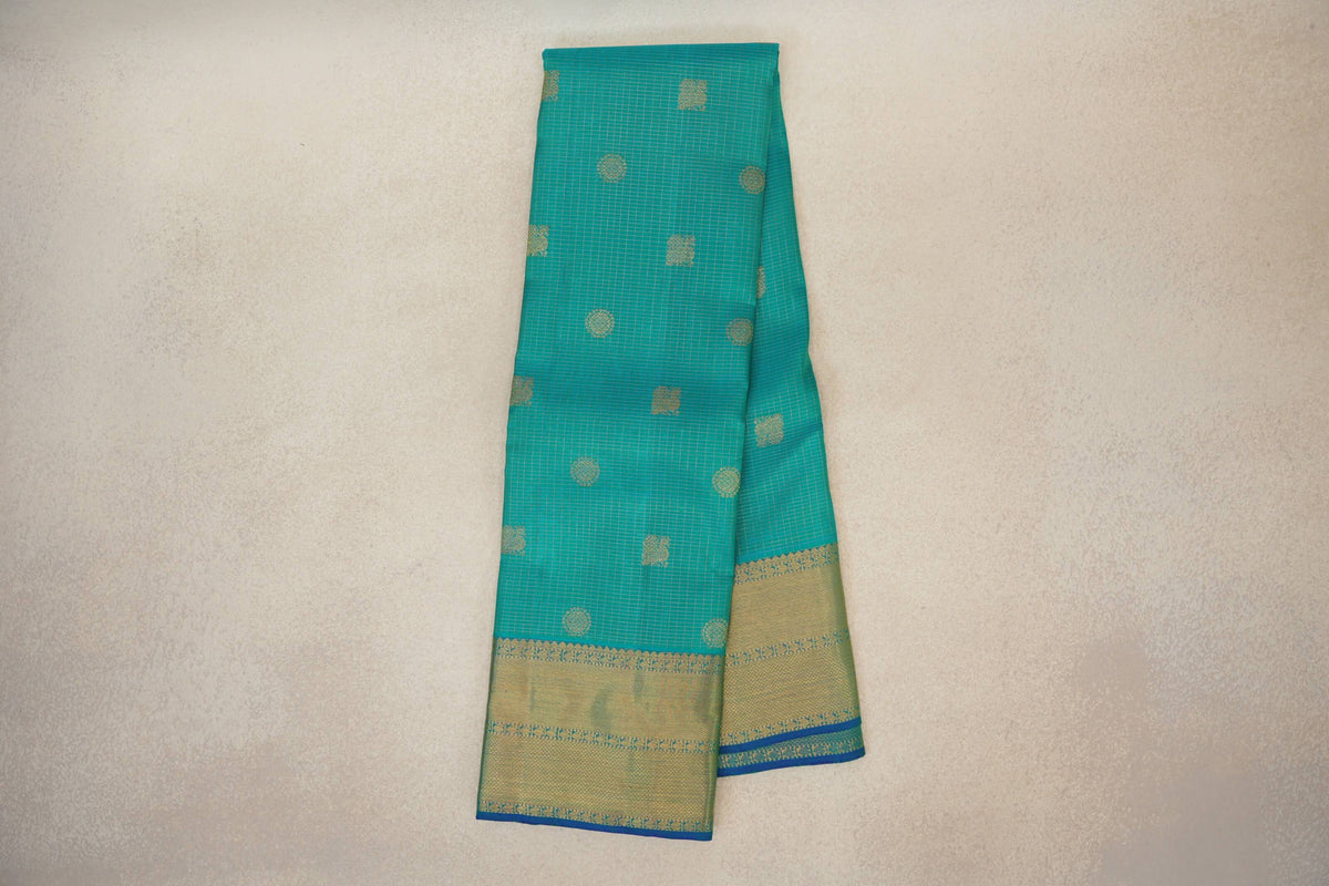 Folded aqua green Kanjivaram silk saree with fine gold checks, scattered circular and square motifs, and broad gold borders edged in blue.