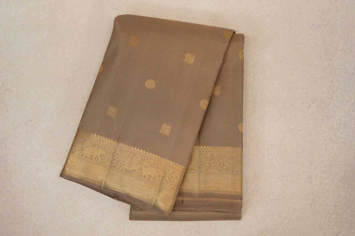Folded greenish beige Kanjivaram silk saree with fine horizontal lines, scattered gold motifs and a wide, intricately patterned gold border.