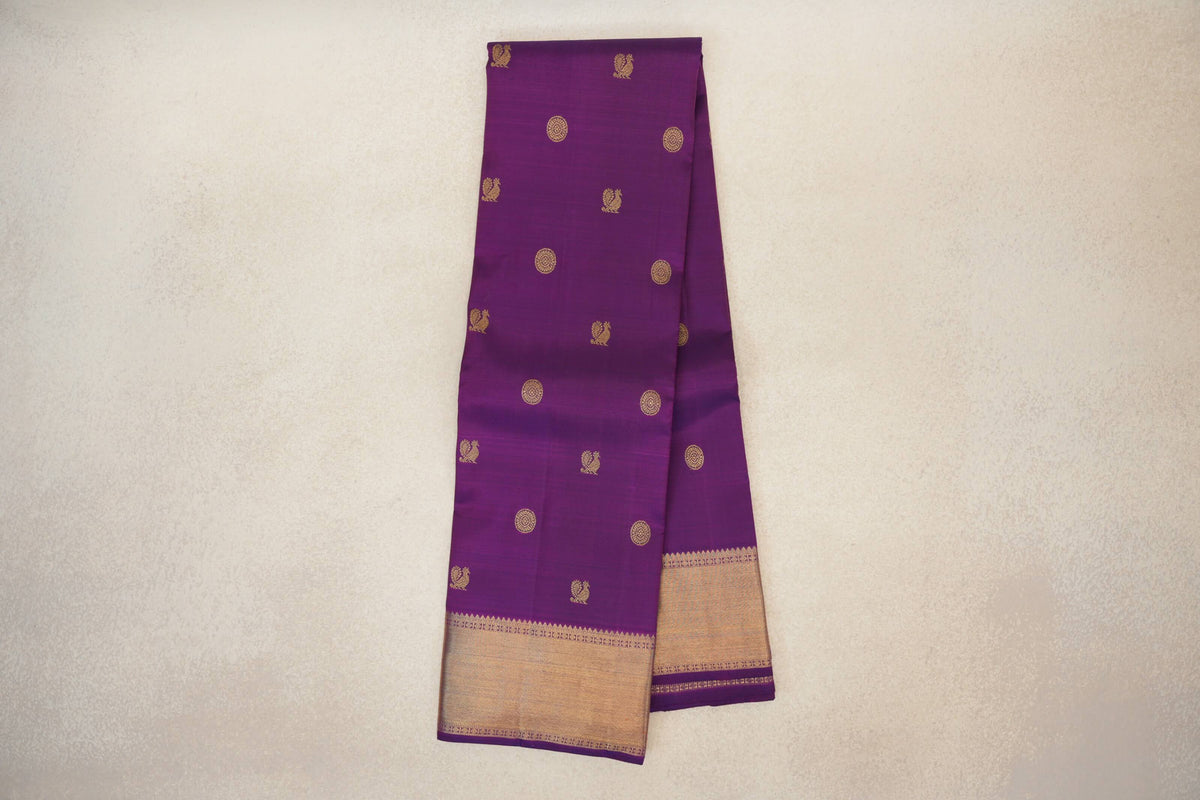 Folded purple Kanjivaram silk saree with scattered gold circular and bird motifs, and a broad gold border along the lower edge.
