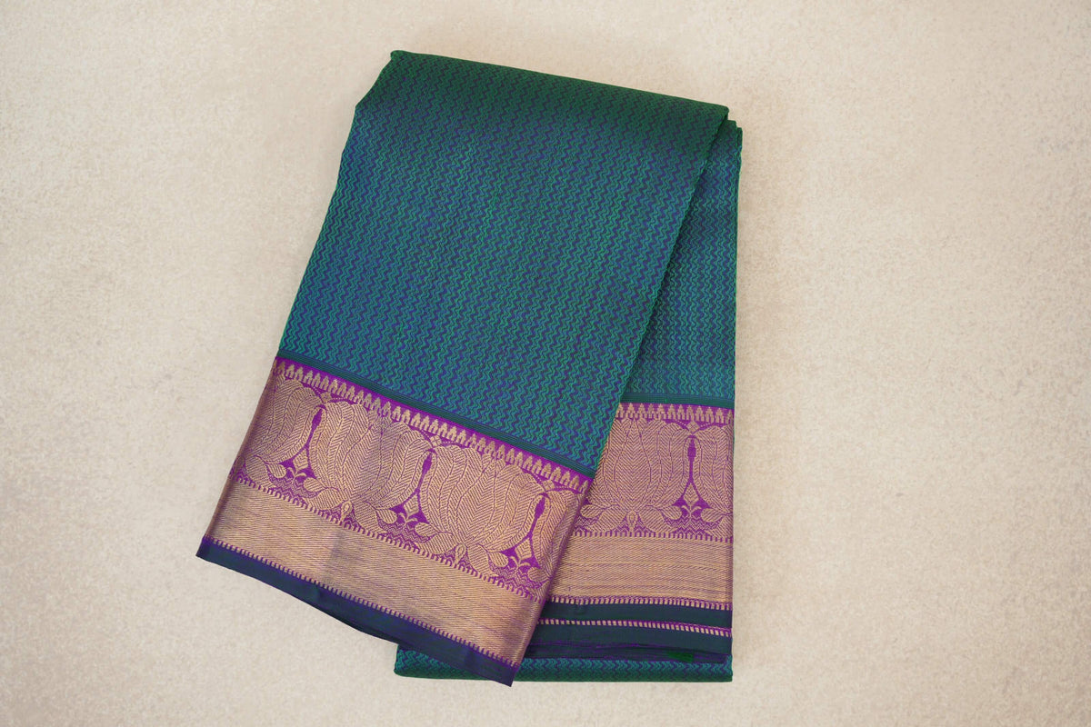 Folded peacock green Kanjivaram silk saree with a fine zigzag pattern and broad purple borders with gold-toned woven designs.