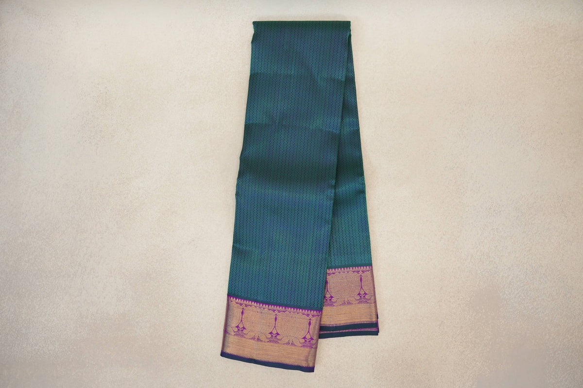 Folded peacock green Kanjivaram silk saree with a fine zigzag pattern and purple borders woven with gold floral motifs and bottle green edging.