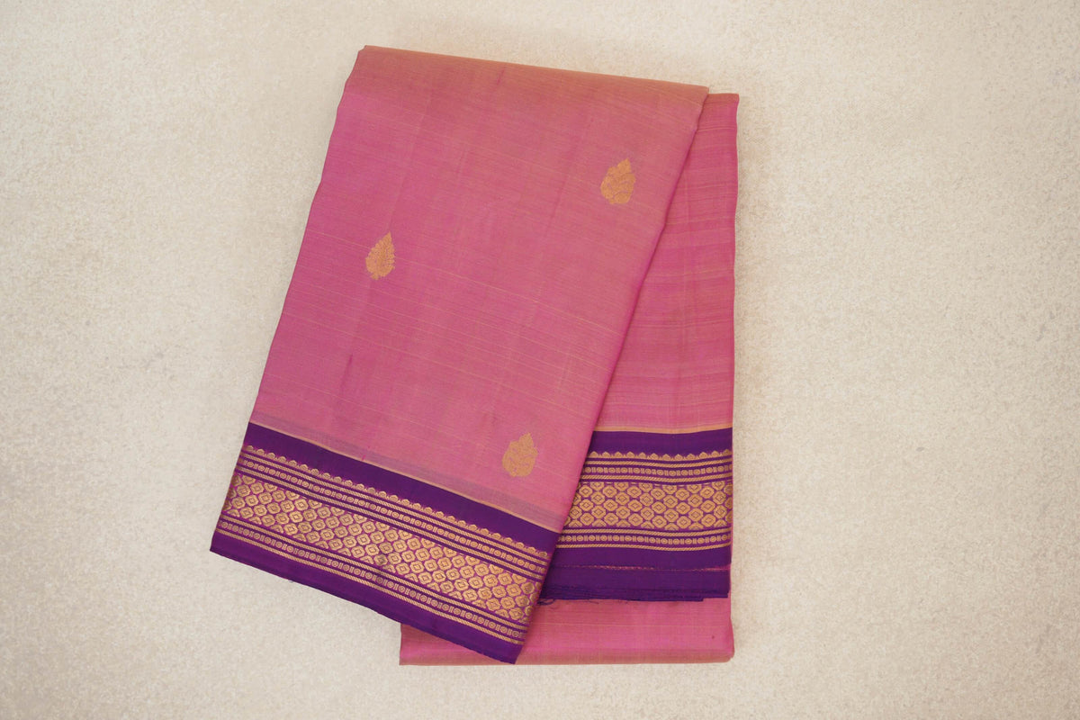 Folded rose pink Kanchipuram silk saree with small gold motifs and a purple border patterned with gold geometric bands.